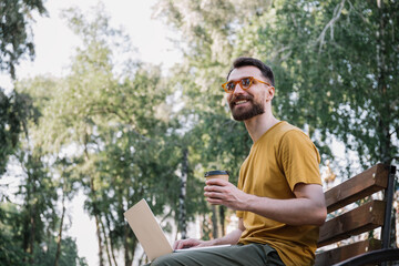 Obraz premium Young freelancer copywriter wearing stylish eyeglasses, working in park, typing on keyboard. Man using laptop computer, holding cup of coffee, shopping online, sitting on bench 
