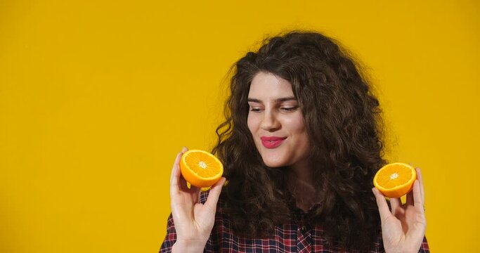 Close-up attractive plus-size model posing. A curly-haired brunette on bright yellow studio background demonstrating different situations. Smiling Girl holding halves of an orange looking at them