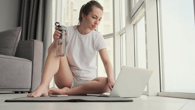 Smiling attractive brunette woman using laptop computer while sitting with bottle on the floor near the window at home