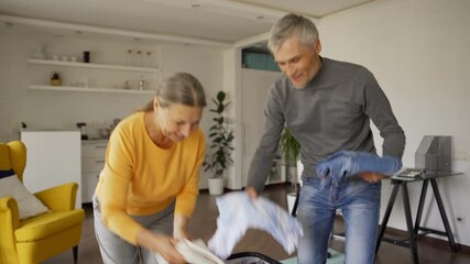 Tilt up medium shot of senior couple entering room with clothes in their hands and packing suitcase for vacations. Happy woman jumping in excitement, trying on straw hat and embracing husband - Powered by Adobe