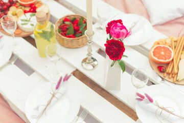A summer celebratory romantic picnic in light pink colors with tasty snacks, fresh fruits and cold lemonade. Floral decorations. Focus on red peonies. Food and drink outdoors