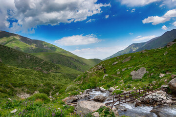 Idyllic summer landscape with hiking trail in the mountains with beautiful fresh green mountain pastures, blue sky, river and brigde.
