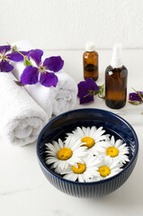 Vertical image.Bowl of chamomile flowers, clean white towels,purple flowers, glass bottles on the marble surface
