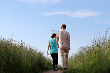 Couple with empty plastic bottle walking through the high grass on background of blue sky and white clouds. Vacation in countryside, going for water, summer travel