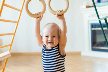 Cute baby performs gymnastic exercises on a wooden home sports complex stairs and rings. Children's...