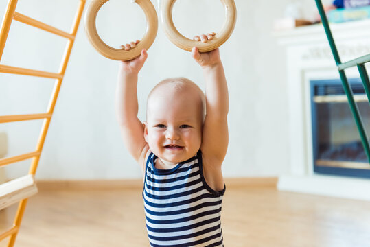 Cute baby performs gymnastic exercises on a wooden home sports complex stairs and rings. Children's sports exercises. Physical education of children at home