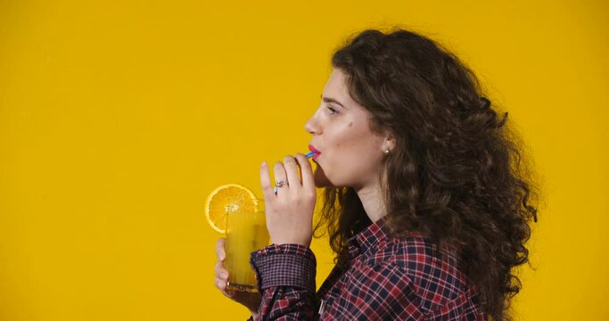 Close-up attractive plus-size model posing. Curly-haired brunette on a bright yellow studio background demonstrating different situations. Girl drinking  cool orange fresh from glass through a straw