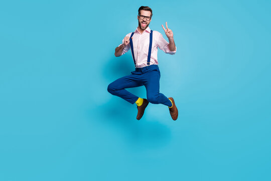 Full length photo of handsome man jump high shopping center crazy addicted shopaholic meet friends show v-sign symbol wear specs shirt suspenders pants boots isolated blue color background