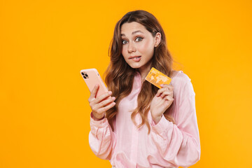 Image of nice confused woman holding credit card and mobile phone