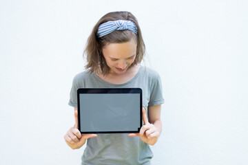 Joyful young woman showing tablet display, looking at blank screen. Front view, person in summer clothes against white background with copy space. Technology and advertising concept