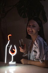 Beautiful girl with a glass of wine on the background of a neon sign of a wine bottle and a glass. Trendy stylish. Glowing life. Modern style. Neon sign.