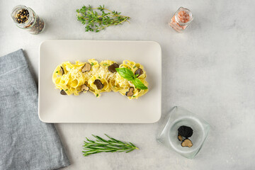 A beige plate with pasta with black truffle and parmesan stands on a grey concrete background. Near are herbs (thyme, rosemary), spices (black pepper, mineral salt) and a bowl with truffle, top view.