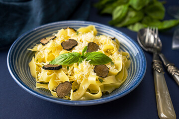 Appetizing homemade fettuccine pasta with black truffle and parmesan in a blue ceramic bowl on a blue background. The dish is decorated with basil leaves. Near are vintage silver cutlery, close-up.