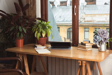 Wooden working desc tabletop with green indoor plants, laptop, books, and notebooks. Window view, rainy autumn weather - obrazy, fototapety, plakaty