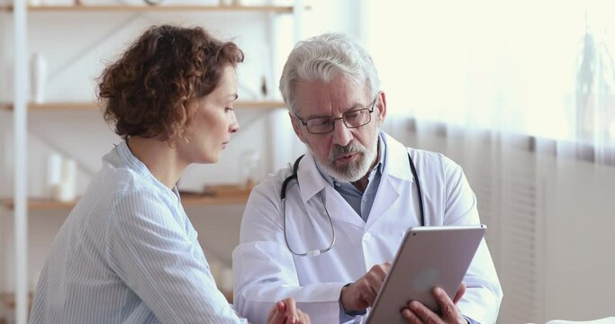 Serious old senior male general practitioner in medical uniform holding digital computer tablet, showing laboratory test results to interested young 30s female patient at checkup meeting in hospital.