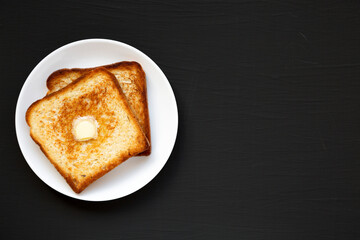 Homemade Buttered Toast on a white plate on a black background, top view. Flat lay, overhead, from above. Copy space.