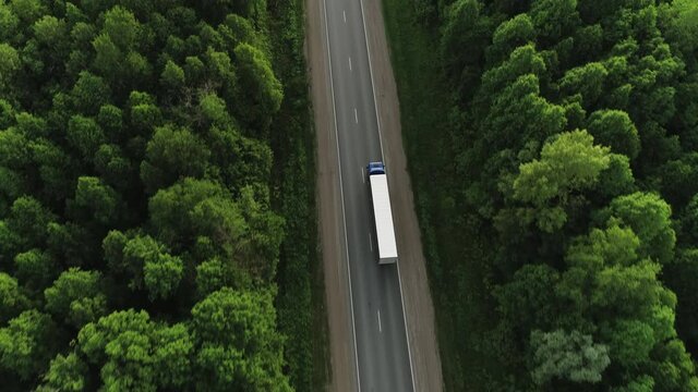 One Semi Truck with white trailer and blue cab driving / traveling alone on dense flat forest asphalt straight empty road, highway top down view follow vehicle aerial footage / Freeway trucks traffic