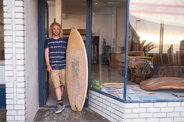 Caucasian male surfboard maker leaning on a door of the entrance holding a new surfboard and smiling