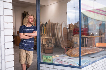 Caucasian male surfboard maker leaning on a door of the entrance with surfboards in the background