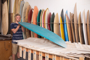 Caucasian male surfboard maker in his studio inspecting one of the surfboards with other surfboards 