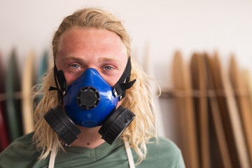 Caucasian male surfboard maker in his studio, wearing a breathing face mask and looking at camera