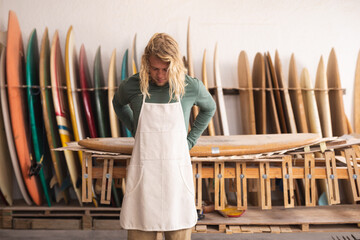 Caucasian male surfboard maker in his studio putting on a protective apron, surfboards in the back