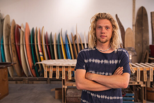 Caucasian male surfboard maker in his studio with surfboards in the background looking at the camera