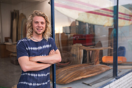 Caucasian male surfboard maker leaning on a door of the entrance with surfboards in the background