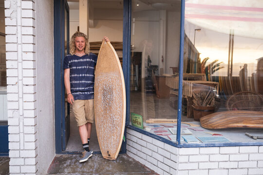Caucasian male surfboard maker leaning on a door of the entrance holding a new surfboard and smiling - Powered by Adobe