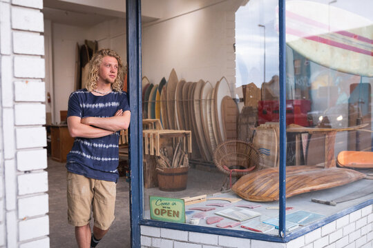 Caucasian male surfboard maker leaning on a door of the entrance with surfboards in the background