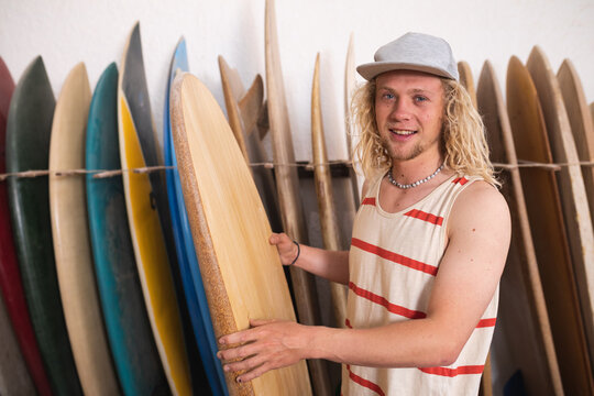 Caucasian male surfboard maker in his studio and holding one of the surfboards and smiling to camera