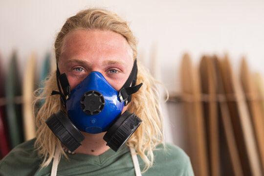 Caucasian male surfboard maker in his studio, wearing a breathing face mask and looking at camera