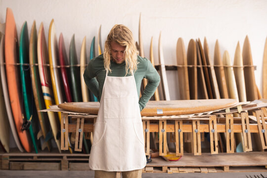 Caucasian male surfboard maker in his studio putting on a protective apron, surfboards in the back