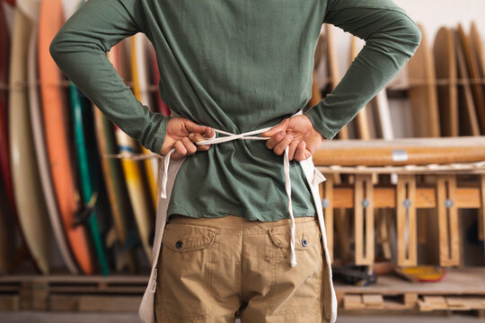 Caucasian male surfboard maker in his studio putting on a protective apron, surfboards in the back - Powered by Adobe