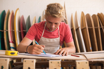 Caucasian male surfboard maker rawing surfboards projects in a sketchbook and making a surfboard