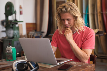 Caucasian male surfboard maker sitting at desk and using his laptop computer with surfboard behind