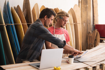 Two Caucasian male surfboard makers working on projects and using a laptop computer with surfboards 
