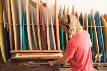Caucasian male surfboard maker wearing protective headphones and cutting wooden stripes