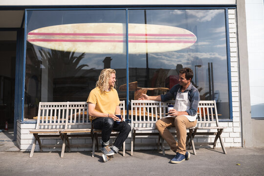 Two Caucasian male surfboard makers sitting on a bench and drinking coffee and having a snack - Powered by Adobe
