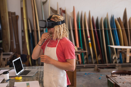 Caucasian male surfboard maker using a VR headset with surfboards in a rack in the background