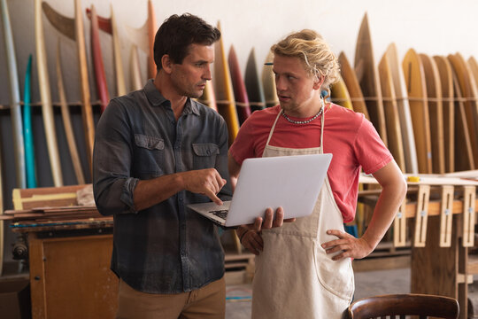 Two Caucasian male surfboard makers working on projects and using a laptop computer