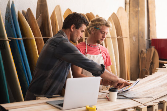 Two Caucasian male surfboard makers working on projects and using a laptop computer with surfboards