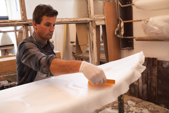Caucasian male surfboard maker preparing a wooden surfboard covered with a white piece of cloth - Powered by Adobe