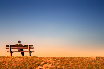 young man resting alone on a bench in an empty landscape at dusk