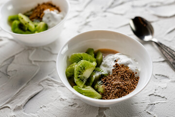 A pair of small dessert bowls with fresh kiwi fruit, white ice cream, yogurt and chocolate chips on a white stone background with teaspoons