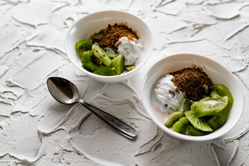 A pair of small dessert bowls with fresh kiwi fruit, white ice cream, yogurt and chocolate chips on a white stone background with teaspoons