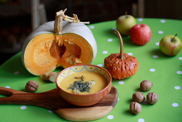 A pumpkin cut in half, pumpkin soup with pumpkin seeds on a wooden cutting board, apples and walnuts on the table