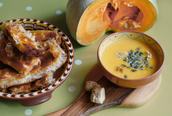 A pumpkin cut in half, pumpkin soup with pumpkin seeds on a wooden cutting board, ginger and walnuts on the table