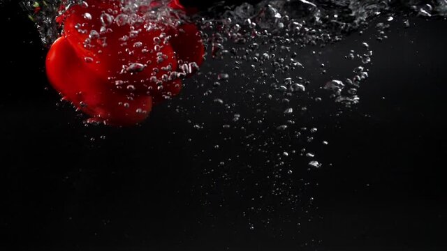 a large fresh red bell pepper falls into the water with splashes, slowly sinking to the bottom, leaving behind a lot of air bubbles. studio shot on a black background. rapid 180 fps