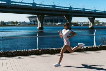 Young fitness woman runner running in the morning on the promenade or embankment and doing sports exercises. Healthy lifestyle, street training, social distancing